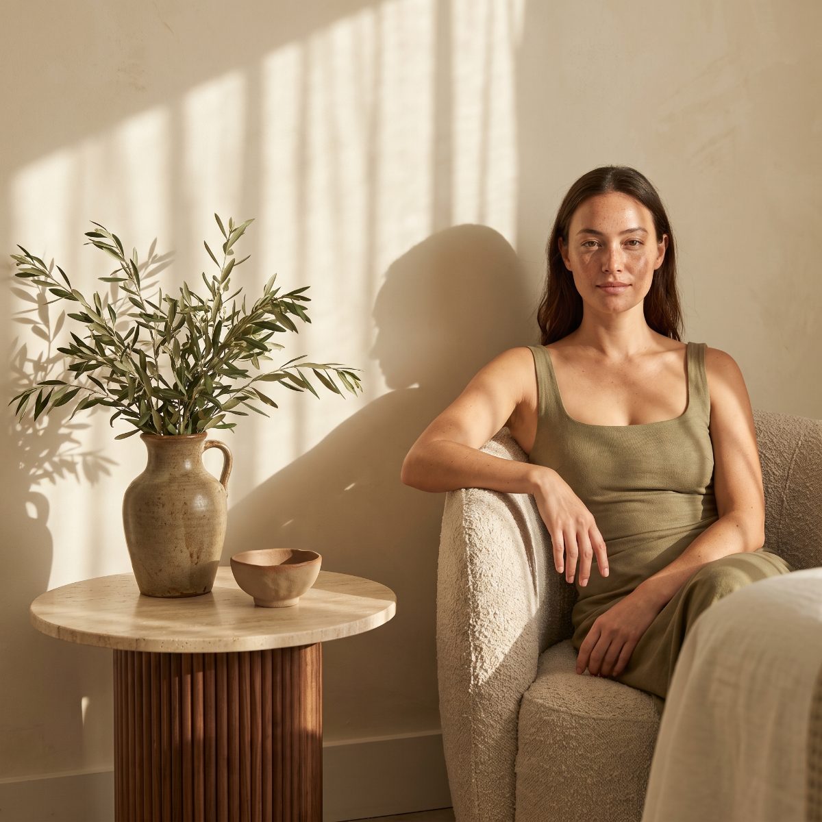 Treatment room corner with an oatmeal armchair, travertine table and olive branches in a vase